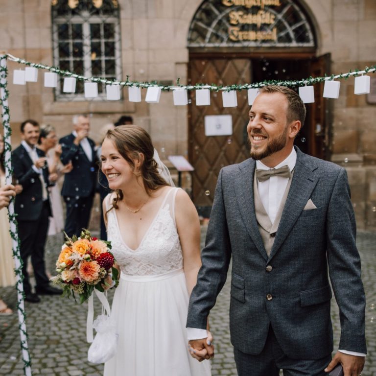 Lachendes Hochzeitspaar unter Konfettiregen und Girlanden, fröhlicher Moment, festliche Atmosphäre | Hochzeitsfotografie – Kathy Audrey Photography.