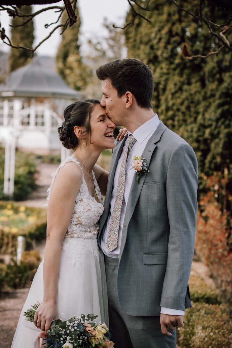 Lachende Braut, Bräutigam küsst ihre Stirn, glücklicher Moment im grünen Garten, Liebe und Freude| Wedding-Photography by Kathy Audrey Photography.