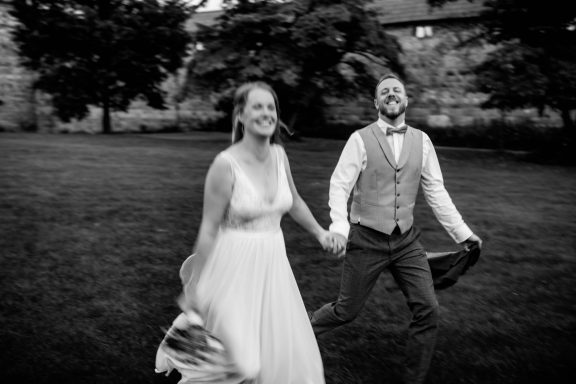 Fröhliches Hochzeitspaar in Schwarz-Weiß, rennend mit natürlichem unscharfem Effekt, voller Lachen und Freude - Kathy Audrey Photography.