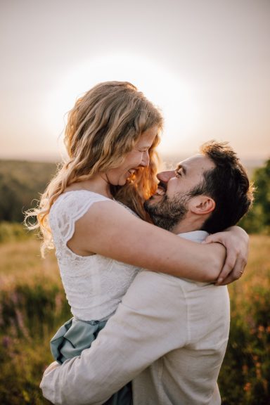 Glückliches Paar im Sonnenuntergang: Er hebt sie hoch, während sie lachen, umgeben von weitem Feld. Kathy Audrey Photography.