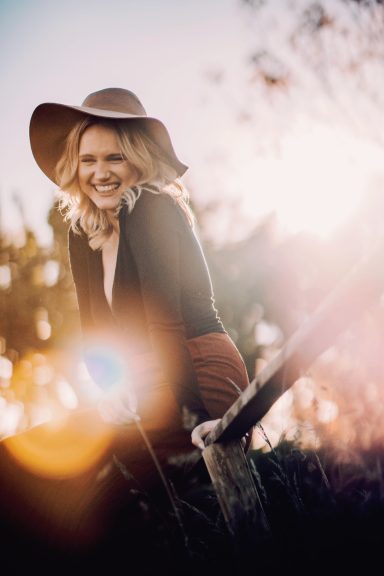 Mädchen mit Hut auf Holzzaun, Feld im Sonnenuntergang, lacht in die Kamera, Kathy Audrey Photography.