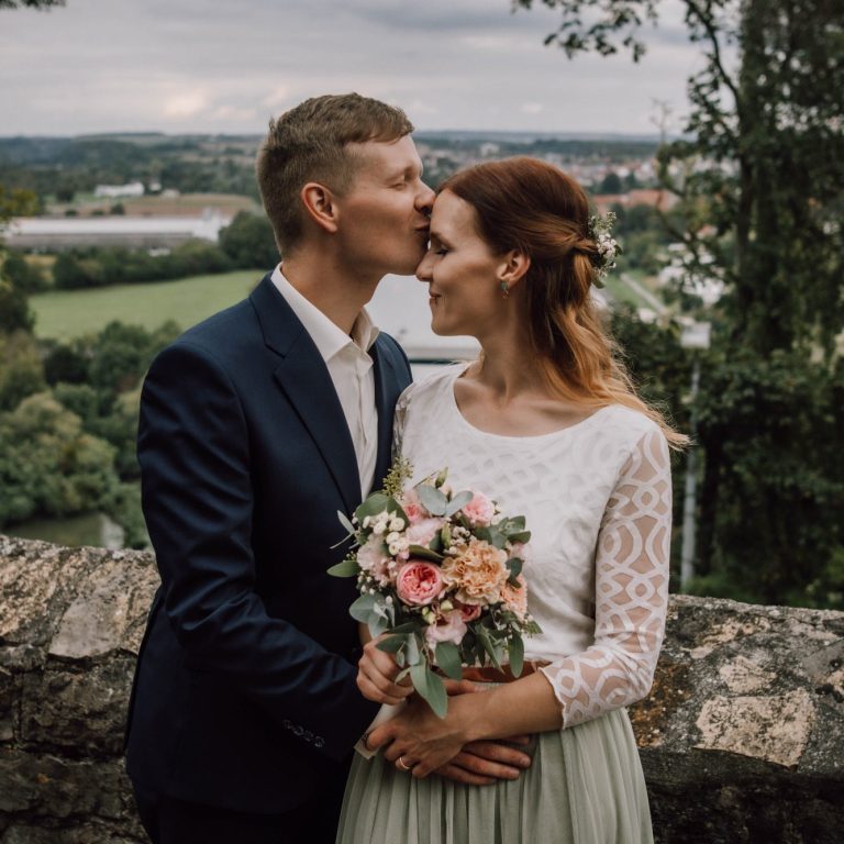 Bräutigam küsst Braut vor romantischer Mauer, Blumenstrauß in der Hand, malerischer Ausblick | Hochzeitsbilder von Kathy Audrey Photography.